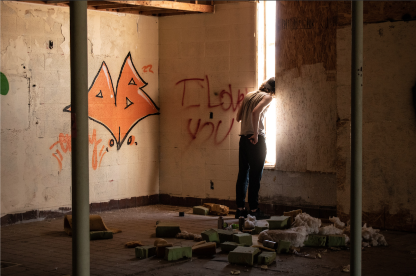 A young man looks out from inside of an abandoned building. His face is obscured by the bright light. The words "I LOVE YOU" are spray painted on the wall next to him.