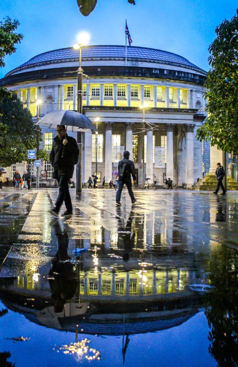 A blue sky behind the illuminated Manchester Library reflected into a puddle while two people walk by, one holding an umbrella which is also reflected in the puddle that takes up the bottom half of the image.
